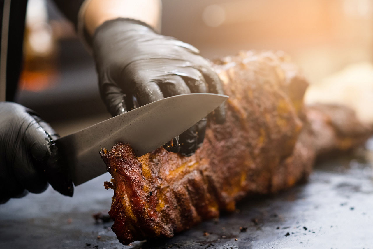 Grill restaurant kitchen. Chef in black cooking gloves using knife to cut smoked pork ribs.