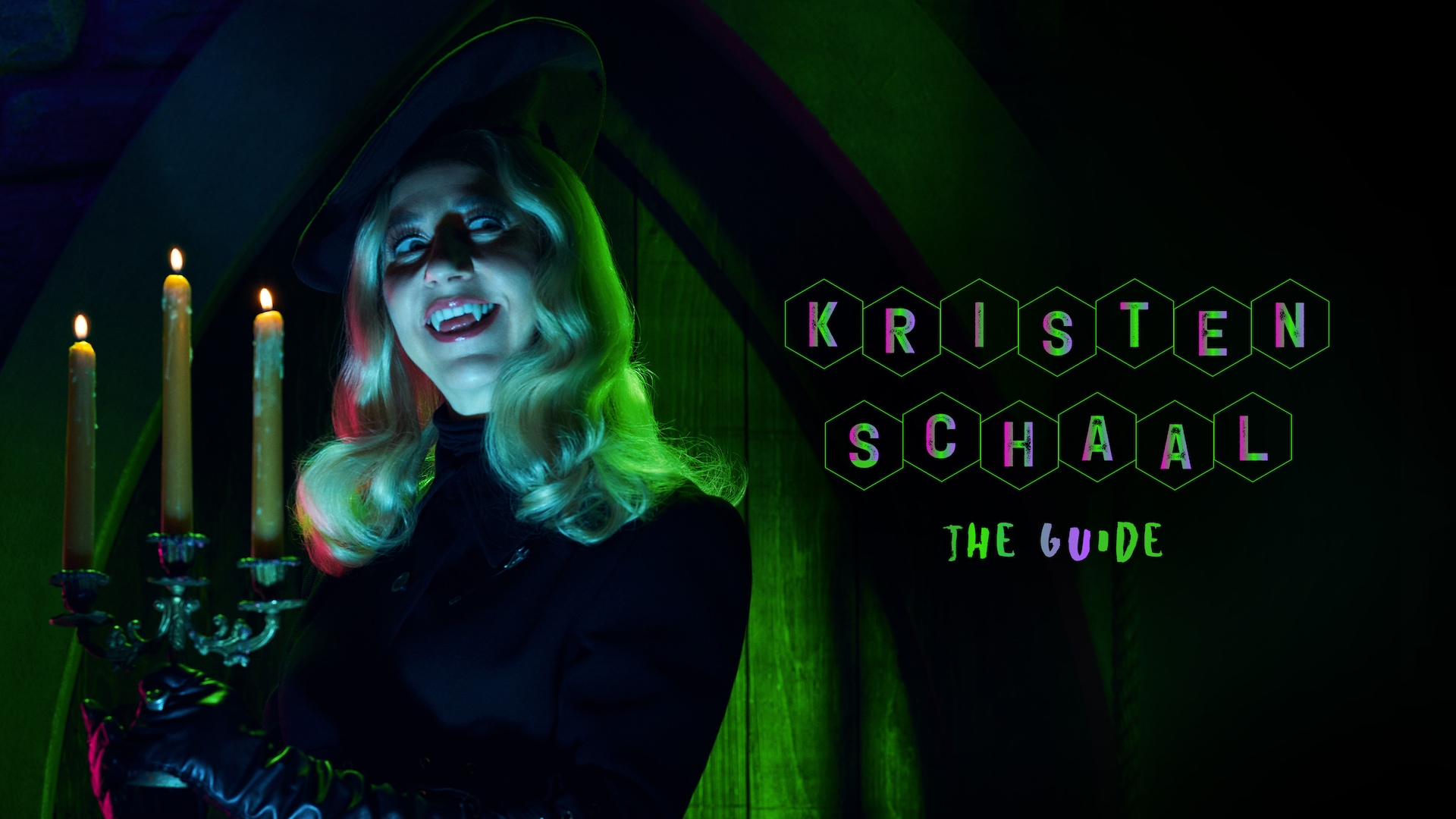 Kristen Schaal as The Guide holding a candelabra, dressed in all black with green and teal lights lighting her face up.