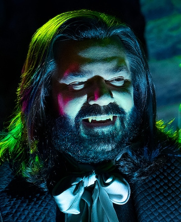 Matt Berry as Laszlo headshot wearing a black jacket and silver ribbon with his face lit with blue, green and pink lights.