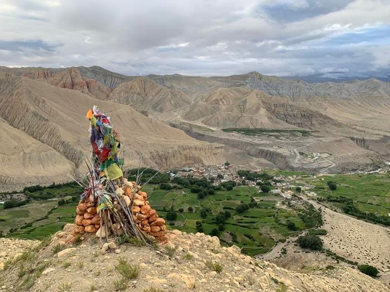 A photo showing a monument made of piled clay-red stones and thin sticks assembled around what looks like different colored flags. The monument is on a hillside looking down on a Tibetan village nestled within green fields among harsh, barren looking hillsides. 