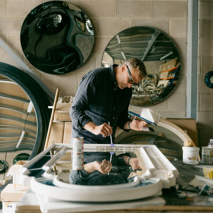 A craftsman paints a frame in a workshop with circular mirrors mounted on brick walls, surrounded by tools, boxes, and materials.