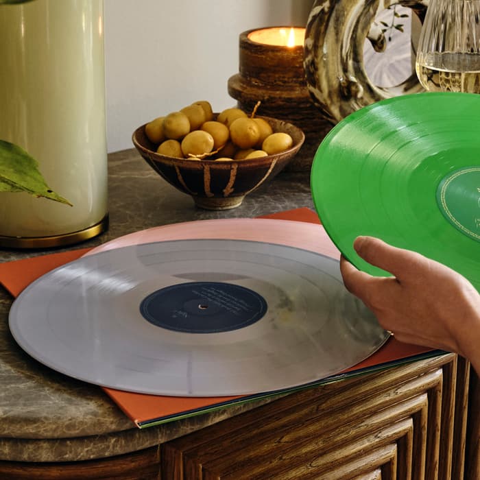 A hand holds a green vinyl record above a table. Another vinyl record lies on the table. Nearby are a candle, a fruit bowl, a plant, and a glass of white wine.