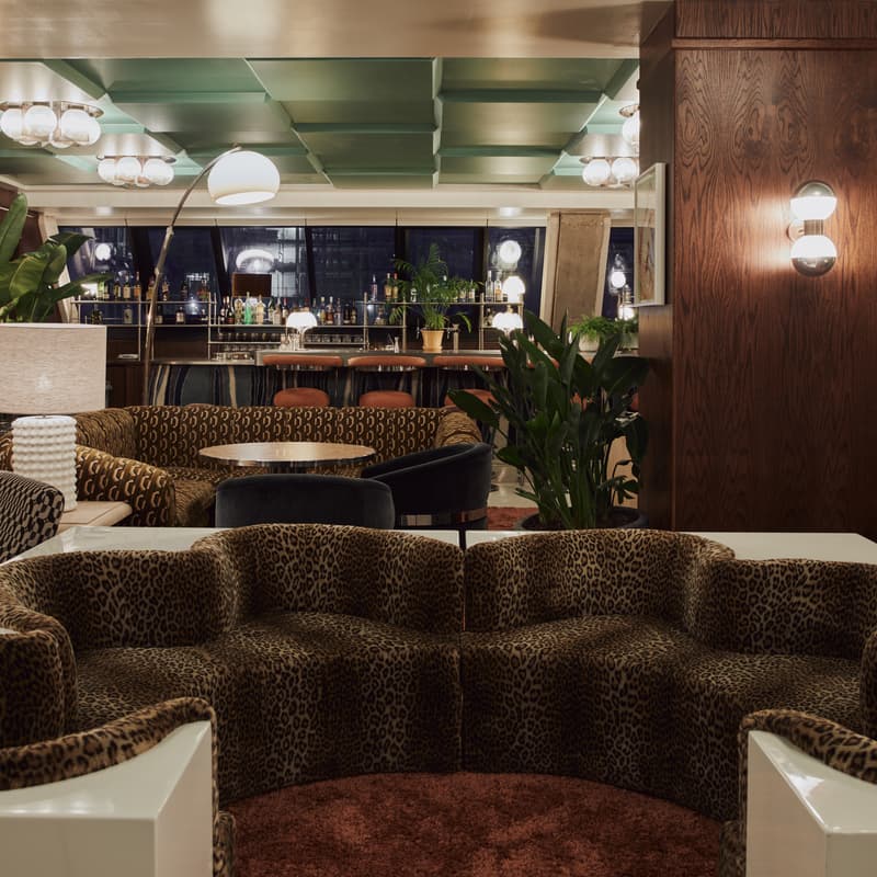 Leopard-print sofa sits in a stylish, dimly lit lounge with patterned chairs, surrounding tables, plants, retro light fixtures, wood accents, and a mirrored wall, leading to a well-stocked bar area.