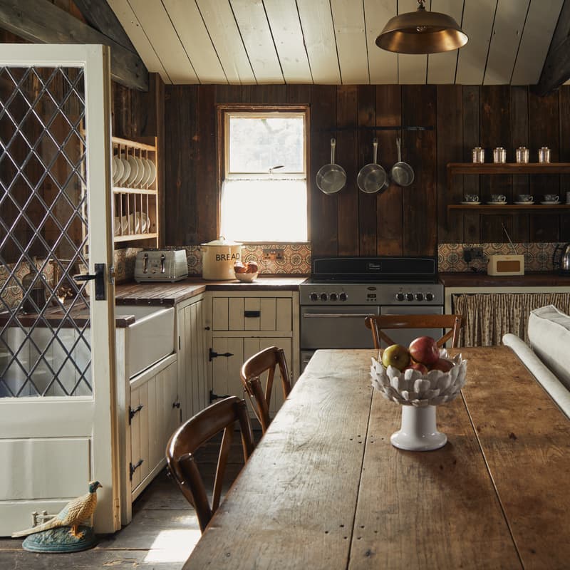 A cozy wooden kitchen with a large wooden table holding a fruit bowl, vintage appliances, open shelves, a window, and rustic décor, including a “BREAD” container on the counter.