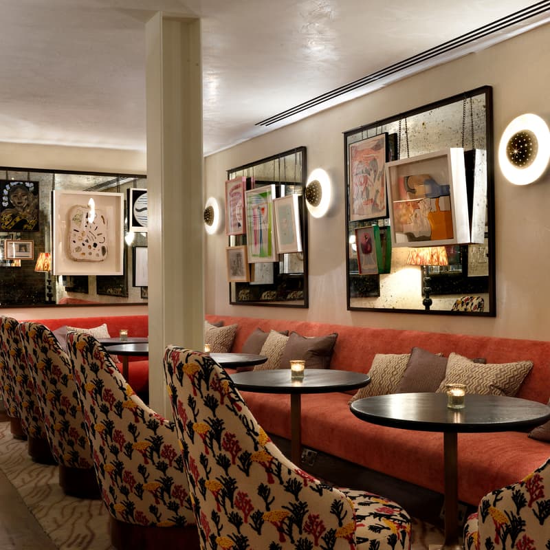 Tables and cushioned chairs with floral upholstery are arranged in a dimly-lit, cozy restaurant. Candle-lit, round tables line a red banquette against an art-adorned wall.