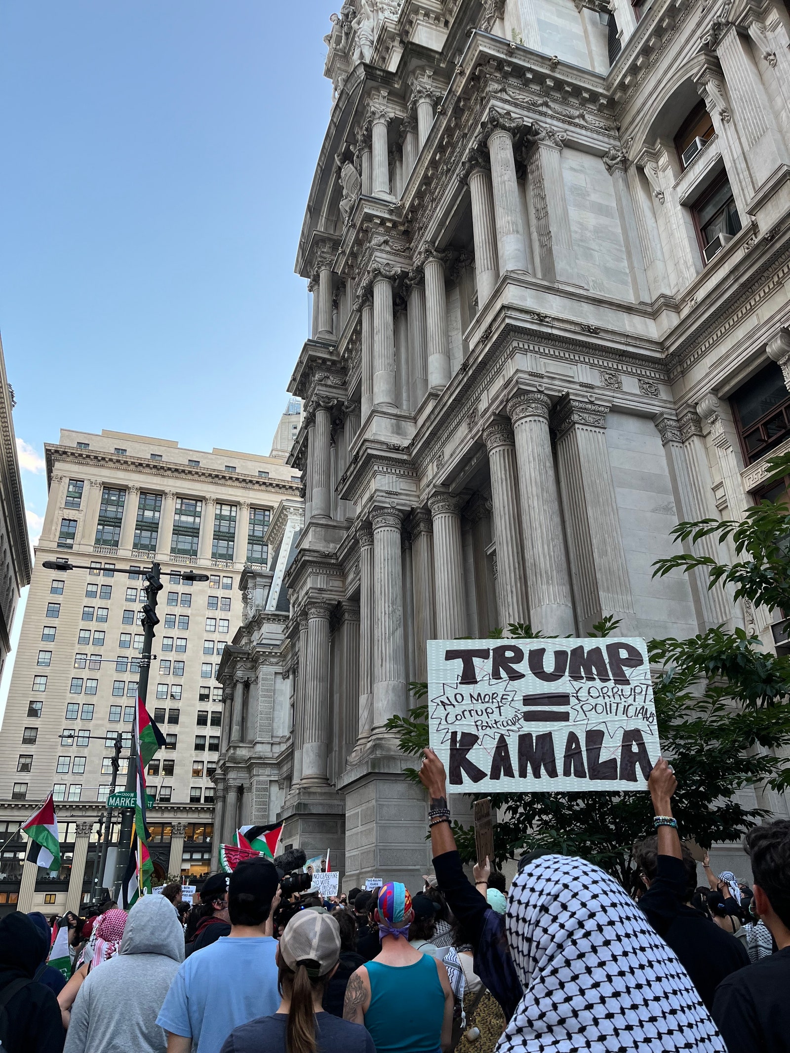 Protesters outside Philadelphia City Hall wait for the march to start. One holds a sign reading “Trump = Kamala No More...