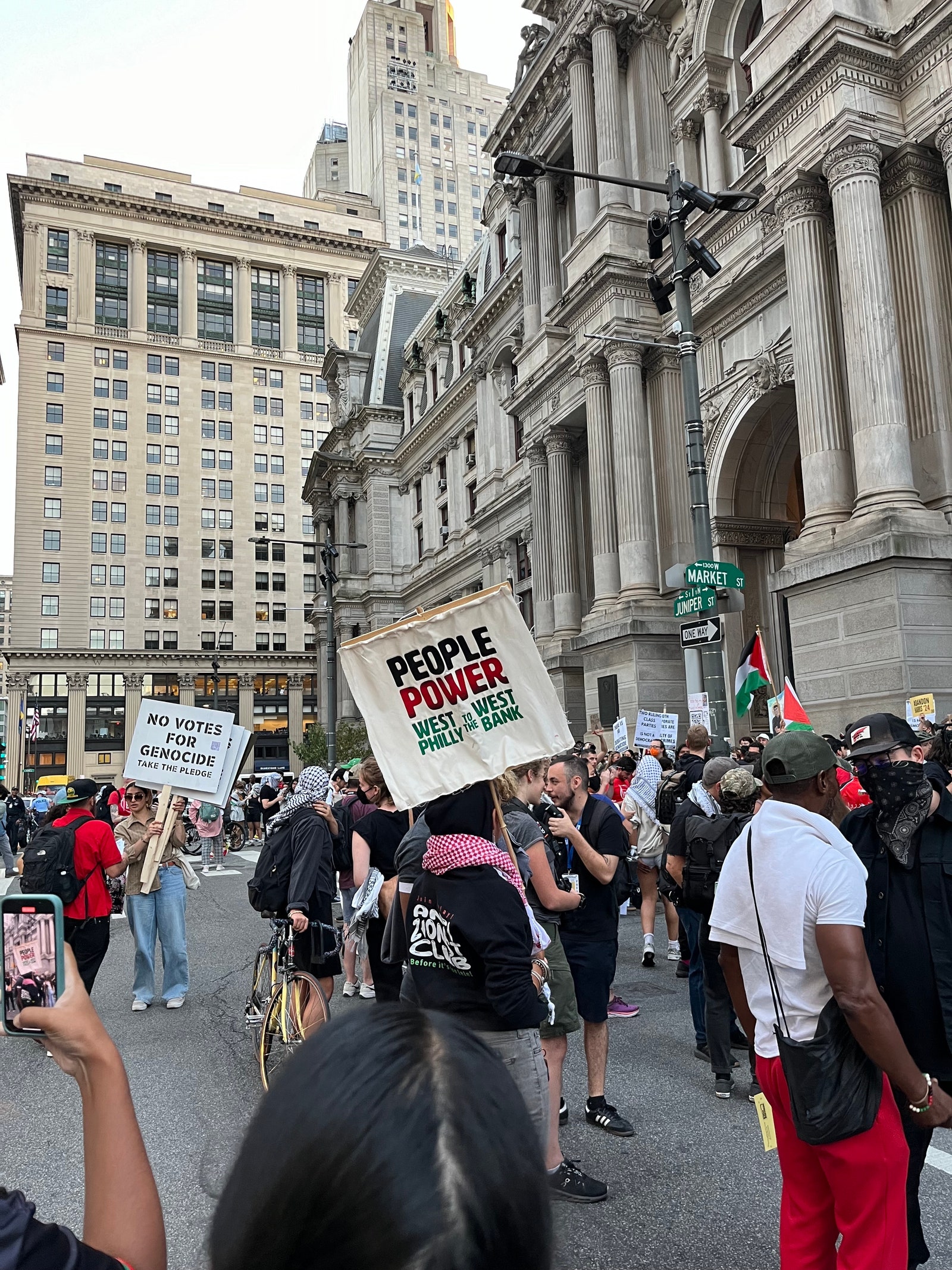 A sign at the Philly debate protest reads “People Power West Philly to the West Bank.” Another behind reads “No Votes...