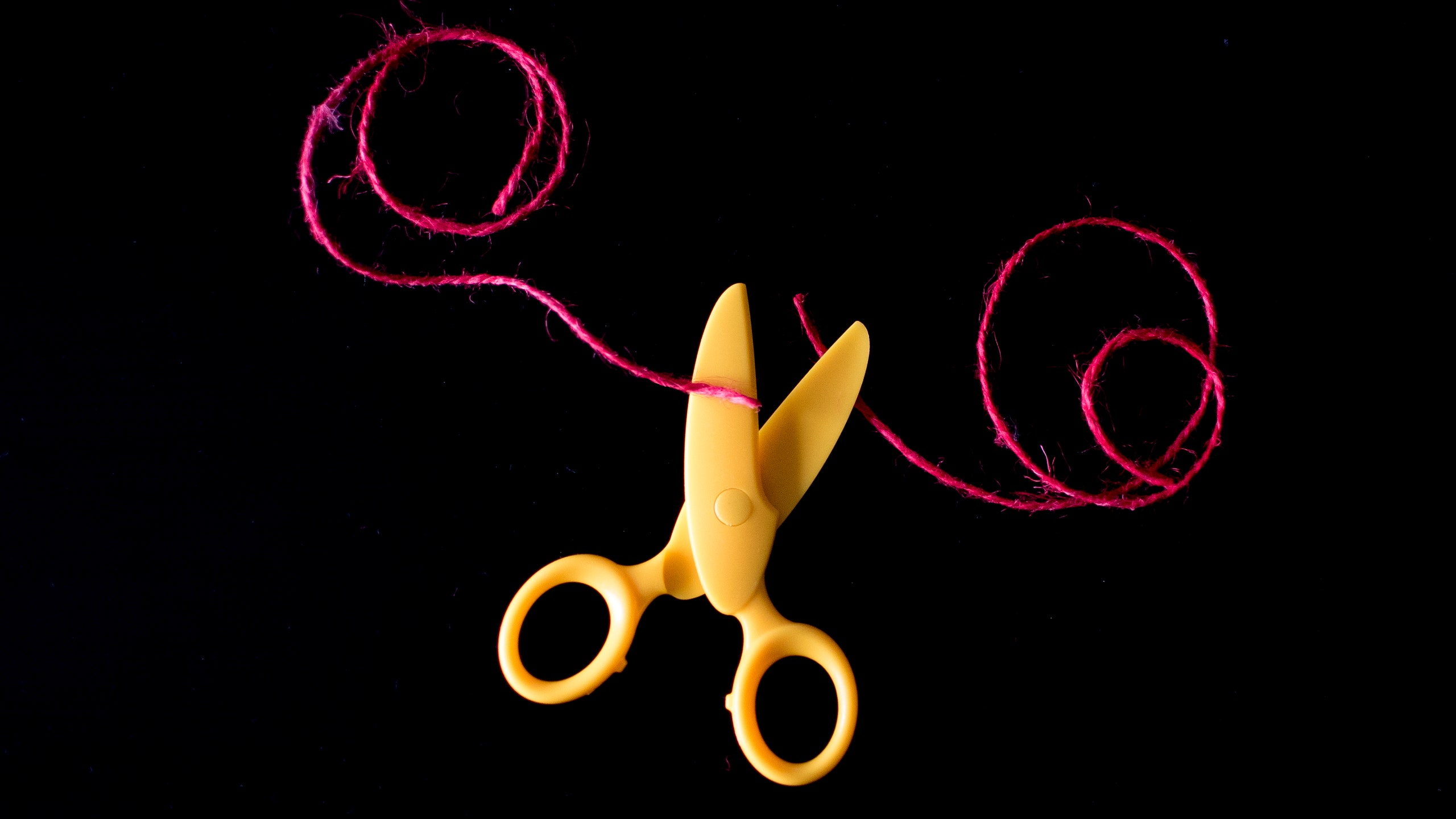 yellow scissors cutting a pink string on a black backdrop.