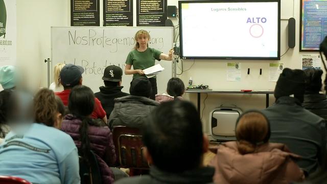 An individual stands in front of a classroom full of people, a white board and a TV monitor. 