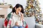 A young woman fell ill during the Christmas and New Year holidays. She sits at home on the sofa covered with a blanket, wipes her nose from a runny nose with a napkin, holds a cup of drink, medicine 