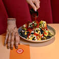 A person slowly pours salsa over a plate of nachos commemorating the hit show 'Wednesday' with inky black tortilla chips. The prop hand from the show stands perched upon its fingertips on the red, diner-style tabletop.