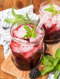 cocktail glass containing blackberries muddled in a bourbon smash on a cutting board with a blue checked napkin