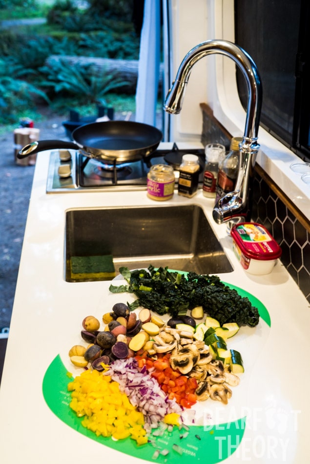 The Kitchen in Kristen's first 4x4 Sprinter camper van showing a dual propane burner, a stainless steel sink and fawcett, and a cutting board with veggies and spices