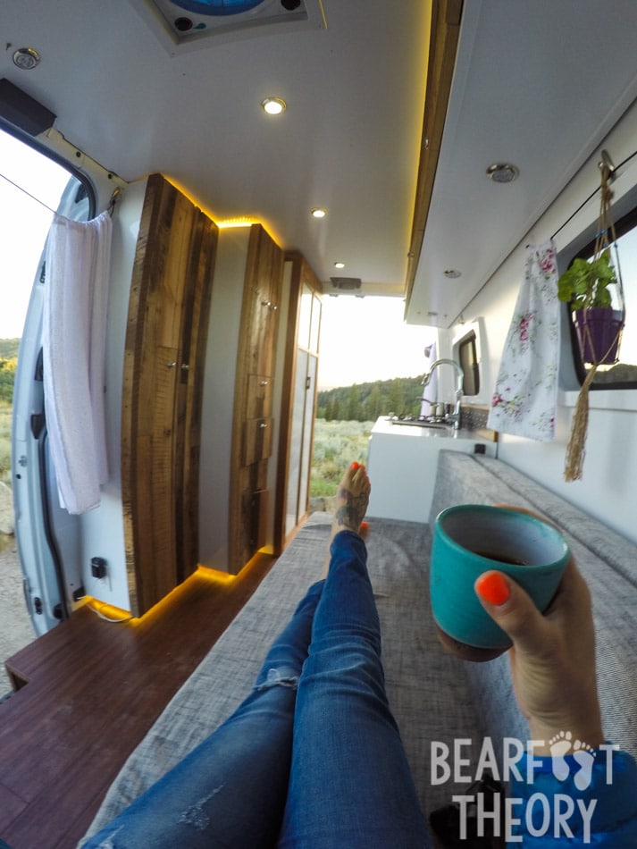 Woman sitting inside her converted Sprinter camper van drinking a cup of tea and looking out the back