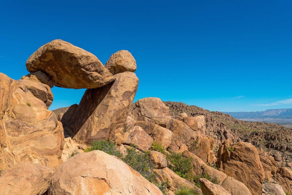 A natural rock formation in a desert landscape, featuring a large, flat boulder curiously perched atop another rock, creating a dramatic arch-like structure. The sky is clear and blue, and the background displays rugged, rocky terrain extending towards distant mountains.