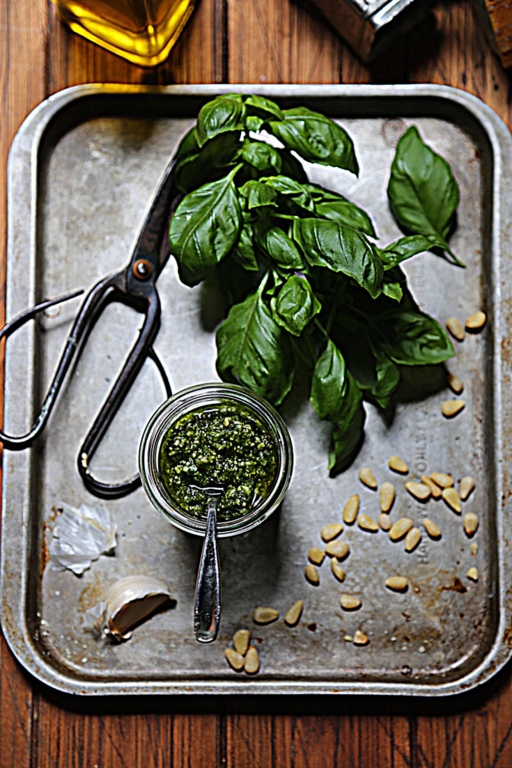 basil, scissors, pine nuts, garlic clove and glass jar of pesto on baking sheet.