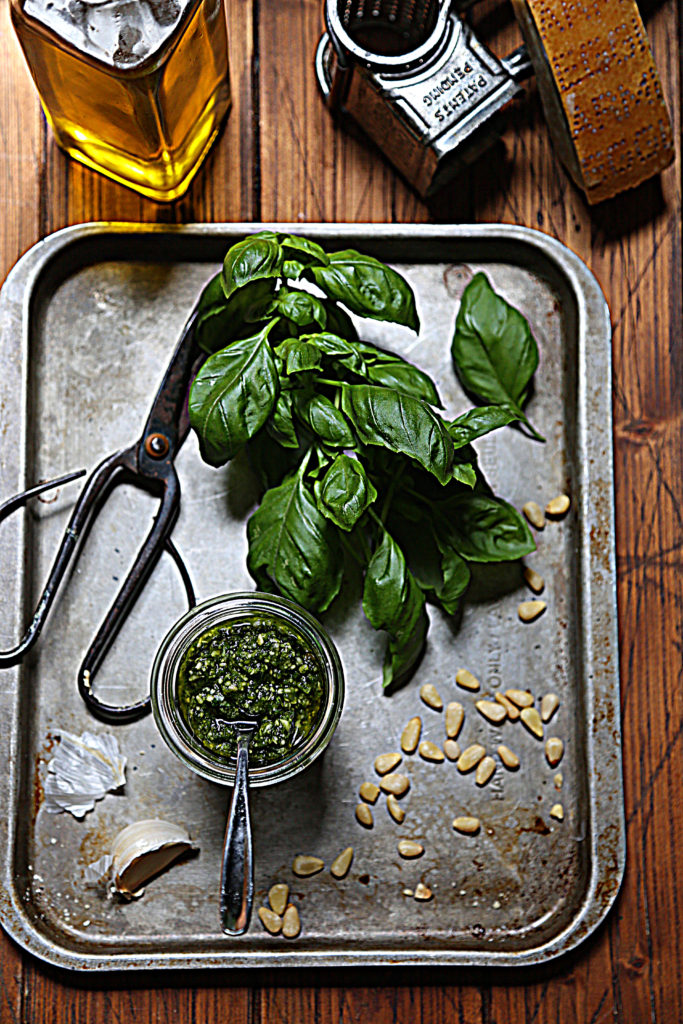 pesto in jar with spoon on baking sheet with basil, pine nuts, scissors and garlic cloves. Glass jar of olive oil and cheese grater in background.