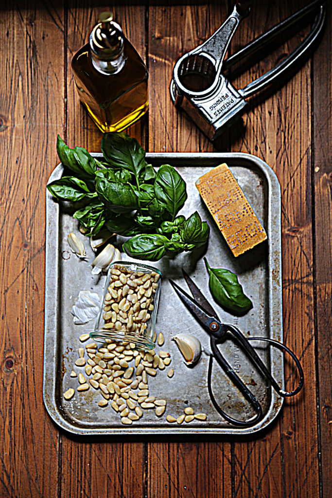 baking sheet with basil, pine nuts in glass jar, garlic cloves, scissors, cheese wedge. Olive oil and cheese grater in background.