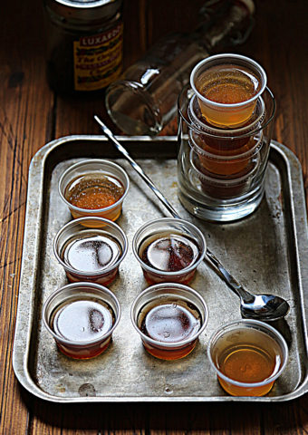 baking sheet with small plastic cups of jello shots. Bar spoon and glass with stacked cups of jello shots.