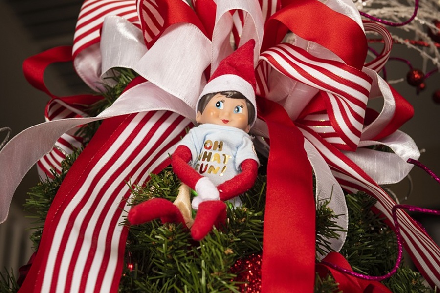 Elf on The Shelf Names an Elf Sitting in a Christmas Tree With a Red and White Ribbon Behind it