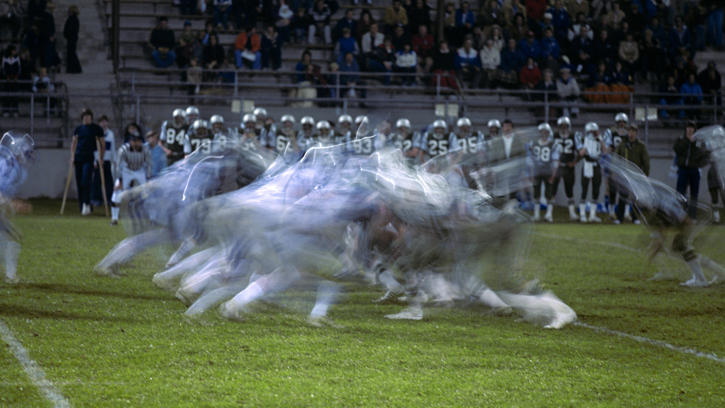 Blurred image of a thrilling football game in action under the night sky, with enthusiastic spectators on bleachers watching one of America's most exciting and sometimes dangerous sports.