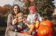 Caucasian mom pushing two toddlers in a wagon with pumpkins with trees in background.