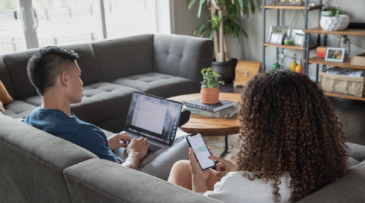 A man and a woman sitting on their couch while looking at their laptop and smartphone.