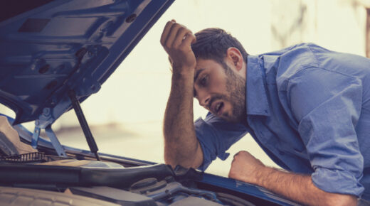 A stressed man leaning over his car’s open hood, looking at the engine.