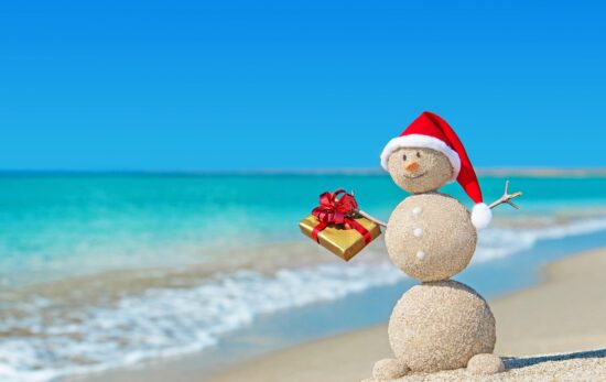 Smiley sandy snowman at beach in christmas hat with golden gift. Holiday concept for New Years Cards.