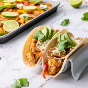 Two soft tortillas filled with Sheet Pan Chicken Fajitas, featuring seasoned chicken, bell peppers, shredded cheese, avocado slices, and cilantro, resting on a taco holder. A sheet pan with more fajita ingredients, lime slices, and fresh cilantro is visible in the background on a marble countertop.