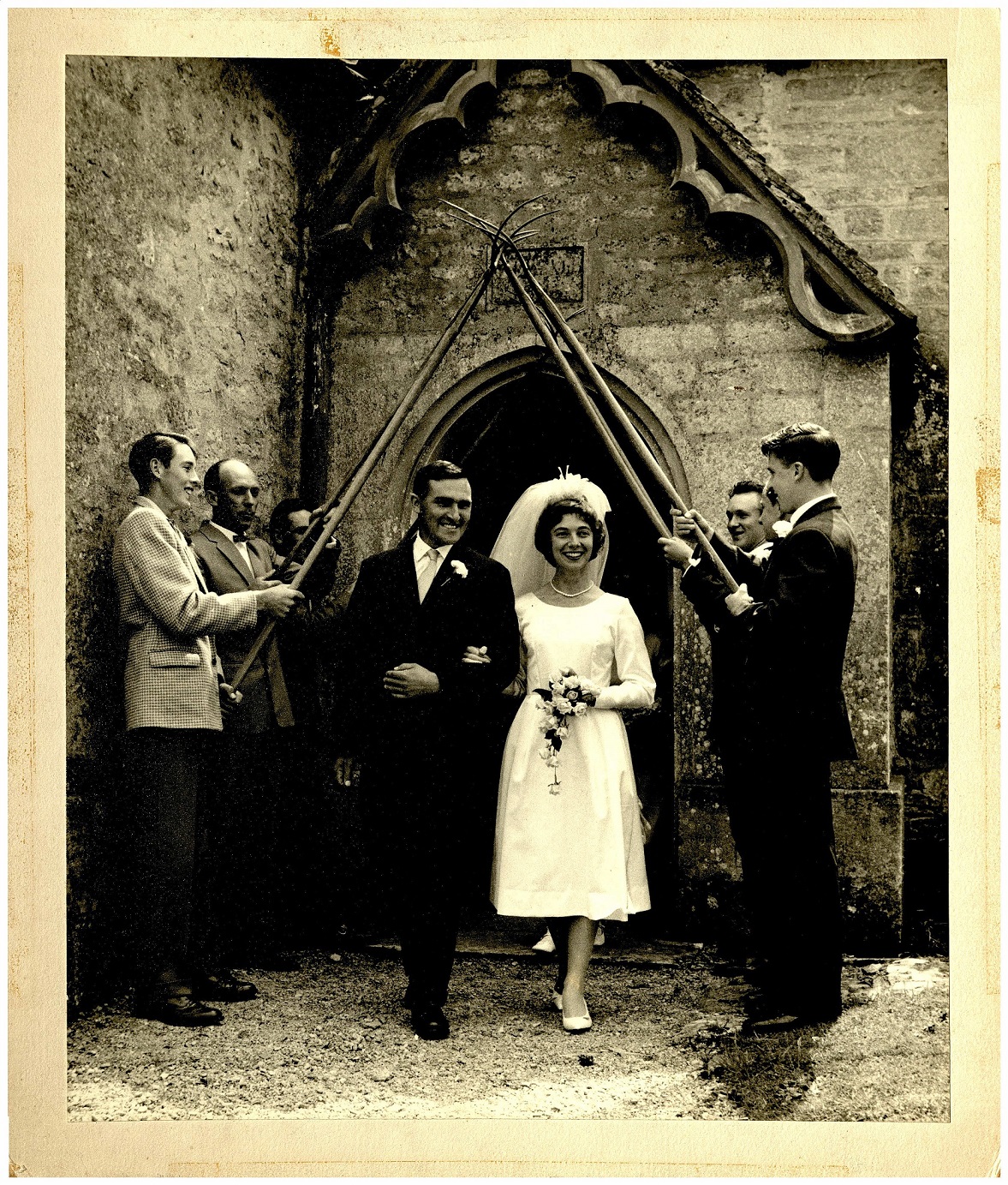 A bridge and groom leaving the church under an arch of pitchforks held up by male wedding guests, 1960s