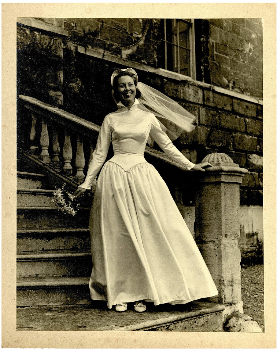 A bride poses on steps wearing a tudor-style wedding dress with a peaked hood and veil, c. 1960s