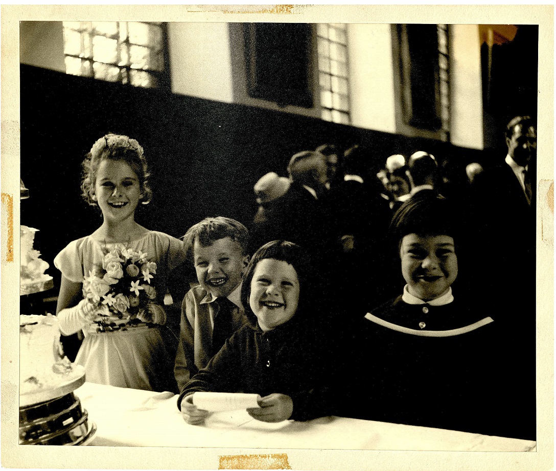 Four children posing by the wedding cake at a wedding reception, one girl dressed as a bridesmaid with flowers, c. 1960s