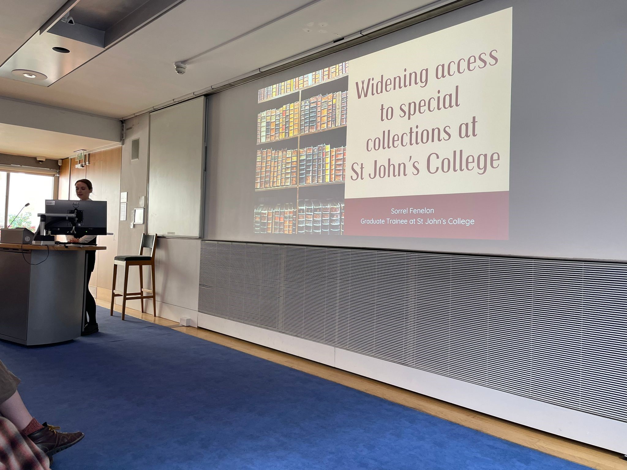 This image shows a powerpoint presentation opened on a screen. The text of the presentation reads Widening Access to Special Collections at St John's College. On the left of the screen there is an a image of some bookshelves with early printed books, with brown covers. On the far left of the image, a woman can be seen standing behind a lecturn presenting. 