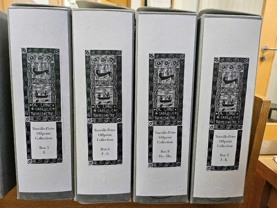 A set of four grey filing boxes. Each box has a white label with a woodprint bookplate which denotes it as part of the Turville-Petre collection. 