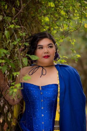 A fat woman stands confidently in a blue corset and matching cape