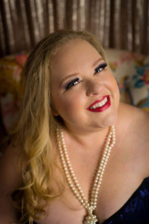 A fat woman wearing a pearl necklace and red lipstick smiles confidently at the camera during a fat-positive photo session