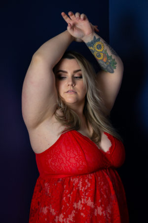 A fat woman in red lingerie holds her arms up with a sultry look to the side during a boudoir photo shoot in the PNW