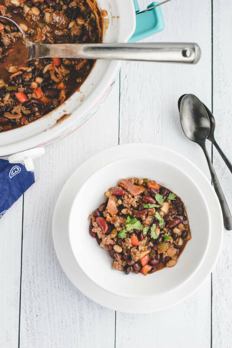 slow cooker with the cowboy beans and white bowl to the side with cowboy beans and black spoons resting at the side.