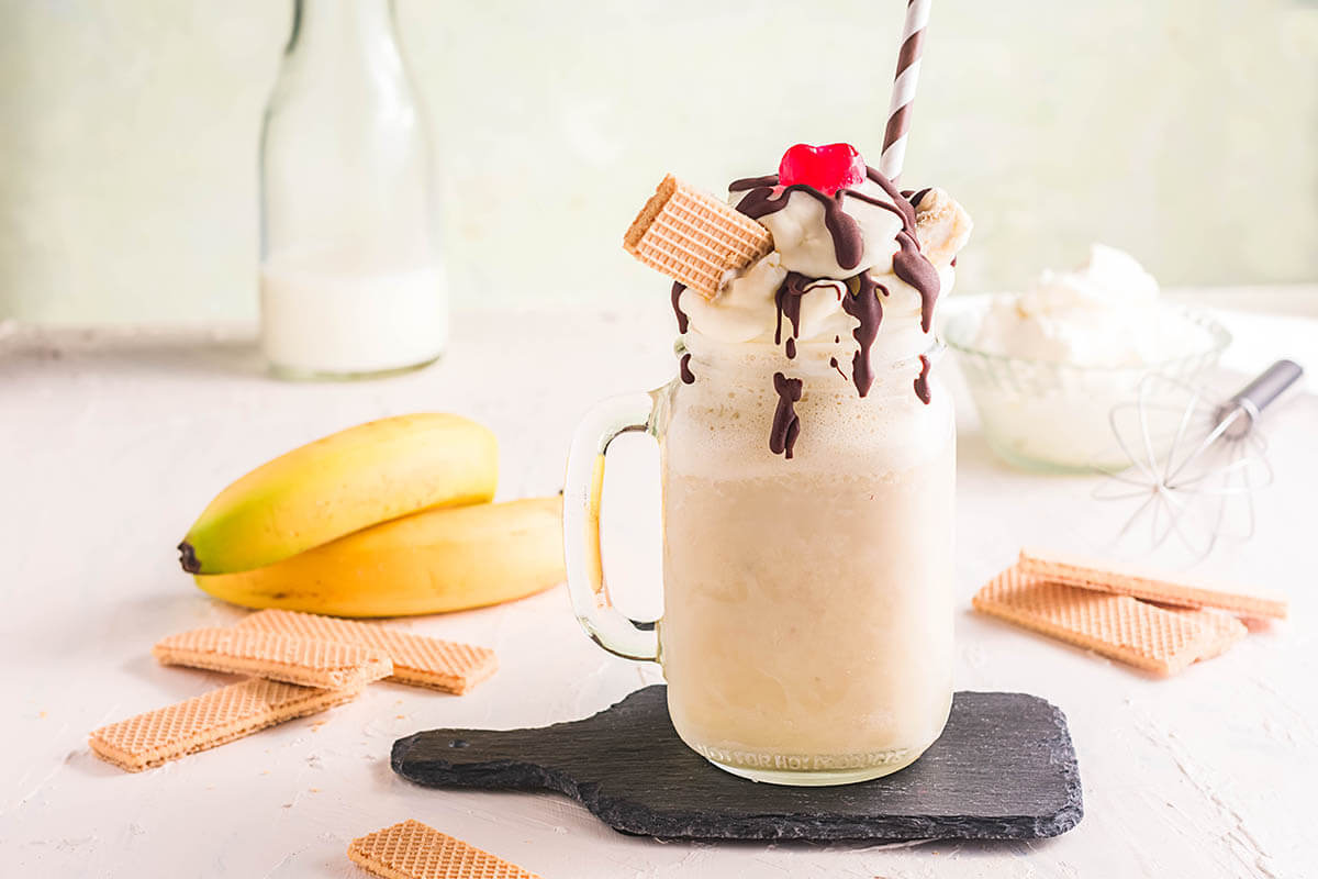 Banana Split Milkshake on table surrounded by bananas, whipped cream and milk.