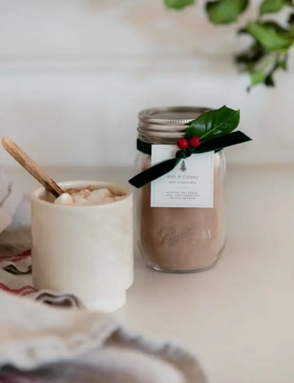 Jar of homemade hot chocolate mix with free printable gift tag and green velvet ribbon on concrete countertop in farmhouse kitchen.