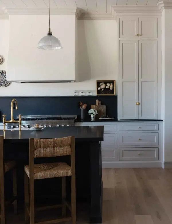 kitchen scene with wicker barstools in front of an all black island, tongue and groove ceiling with plaster hood and light grey cabinets in the background