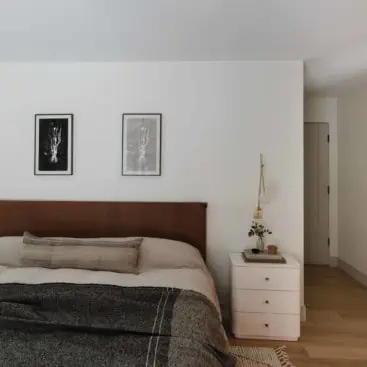 bedroom scene with a carmel eather headboard and footboard, neutral linen bedding with a charcoal throw blanket, black and white art above the headboard. White nightstand with brass and glass modern sconce next to the bed.