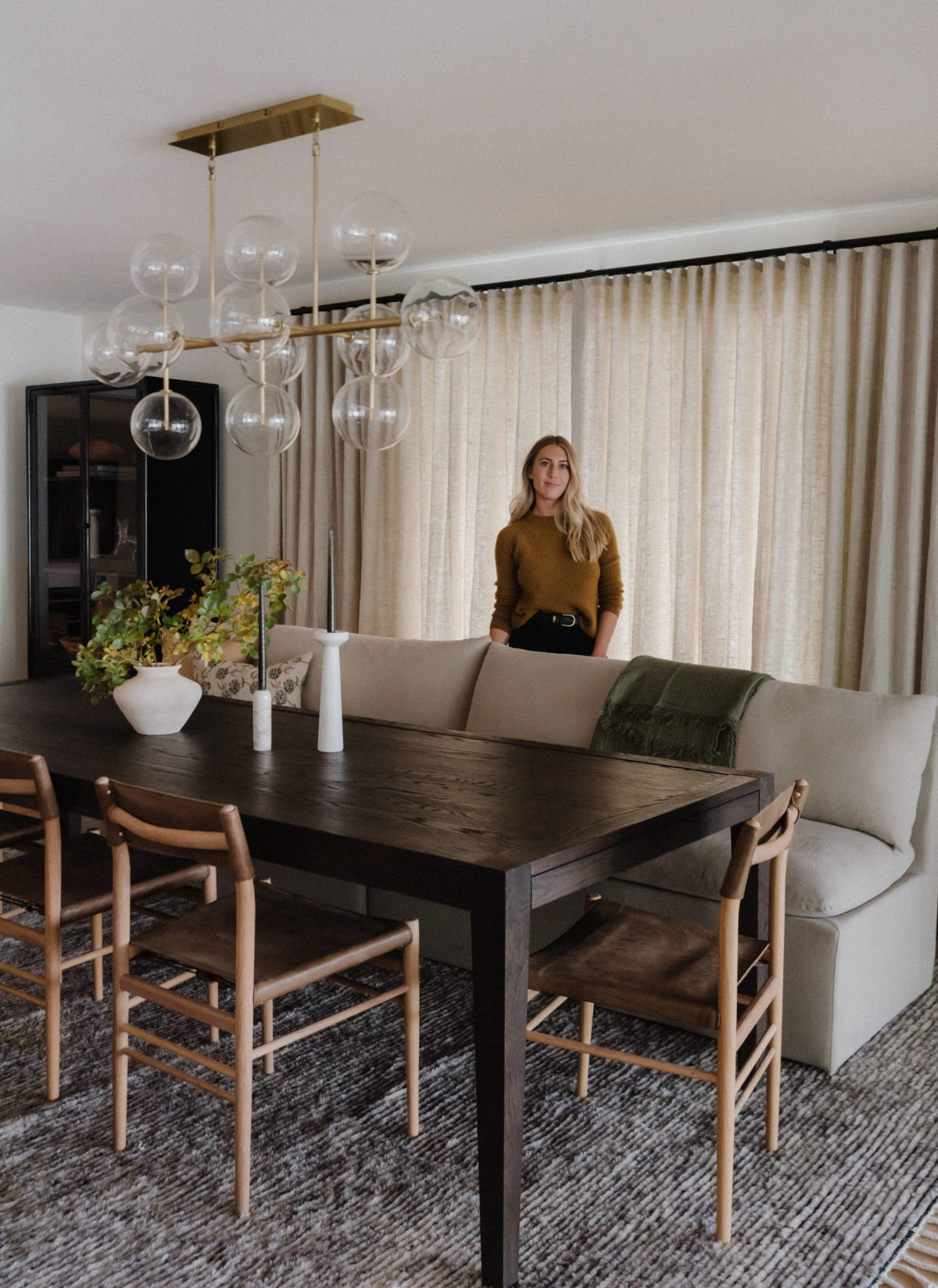 dark wood dining room table with lighter wood chairs on one side with a linen sofa as a bench for the other side. glass and brass bubble light fixture above the table and a woman standing behind the sofa in front of closed linen drapes.