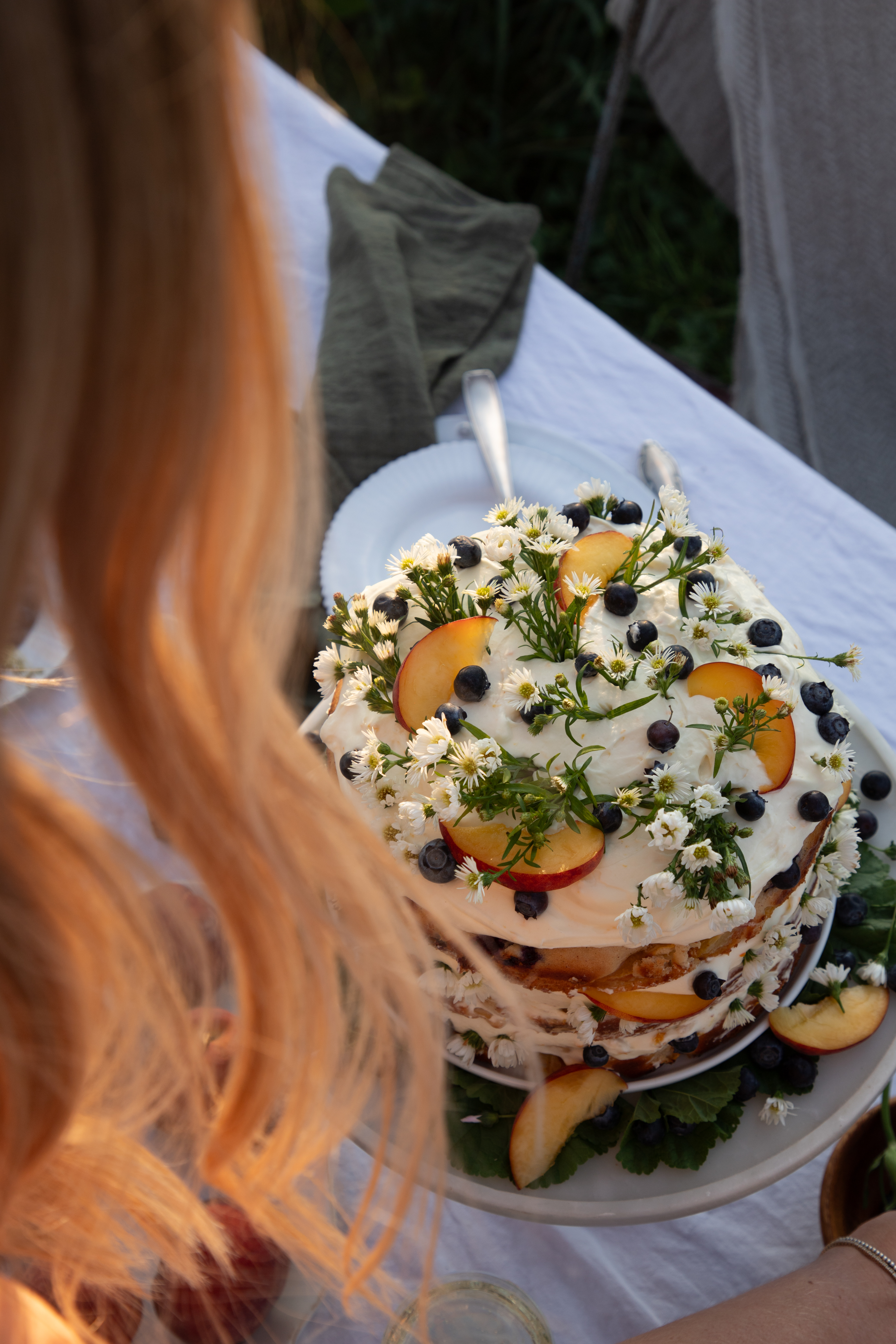 a blonde woman in the foreground looking at a cake covered in camomile flowers, peaches, and blueberries on a marble cake stand.