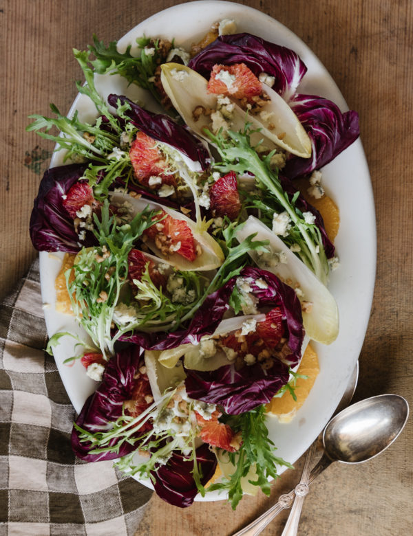 A vibrant endive salad presented on a white plate, featuring crisp Belgian endive and radicchio leaves arranged in a circular pattern. The salad is topped with bright orange slices, crumbled blue cheese, and crunchy walnuts. Fresh parsley adds a touch of green, creating a beautiful and appetizing dish perfect for any gathering.