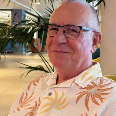 Chris Miller, a man in is 60s, smiling into the camera wearing a bright hawaiian shirt.