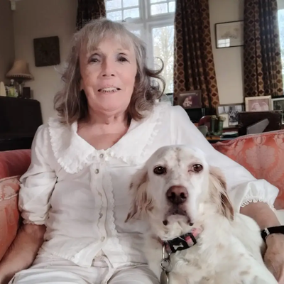 Geraldine, a woman in her 70s, sits at home on a sofa with her dog, Alice, beside her.