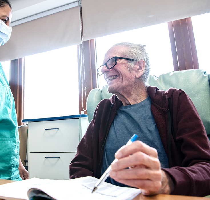 A nurse is caring for a patient she is wearing a blue uniform and holding the patients arm and smiling at him. The patient is smiling back, he is sat down in a chair wearing a checked patterned dressing gown.