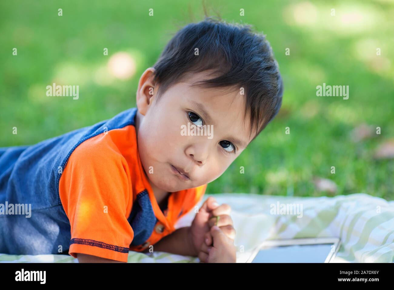 A latino boy taking a break from playing educational games on his tablet, while laying down on the lawn. Stock Photo